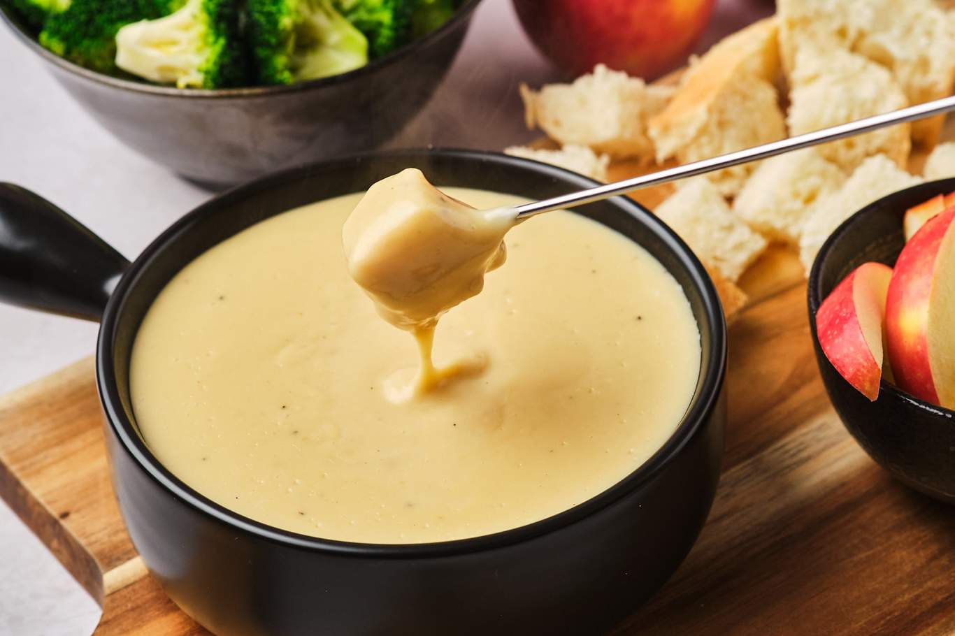 Dipping a cube of French bread into vegan fondue with a fondue fork; the pot of fondue is surrounded by bowls of apples, broccoli, and bread cubes.