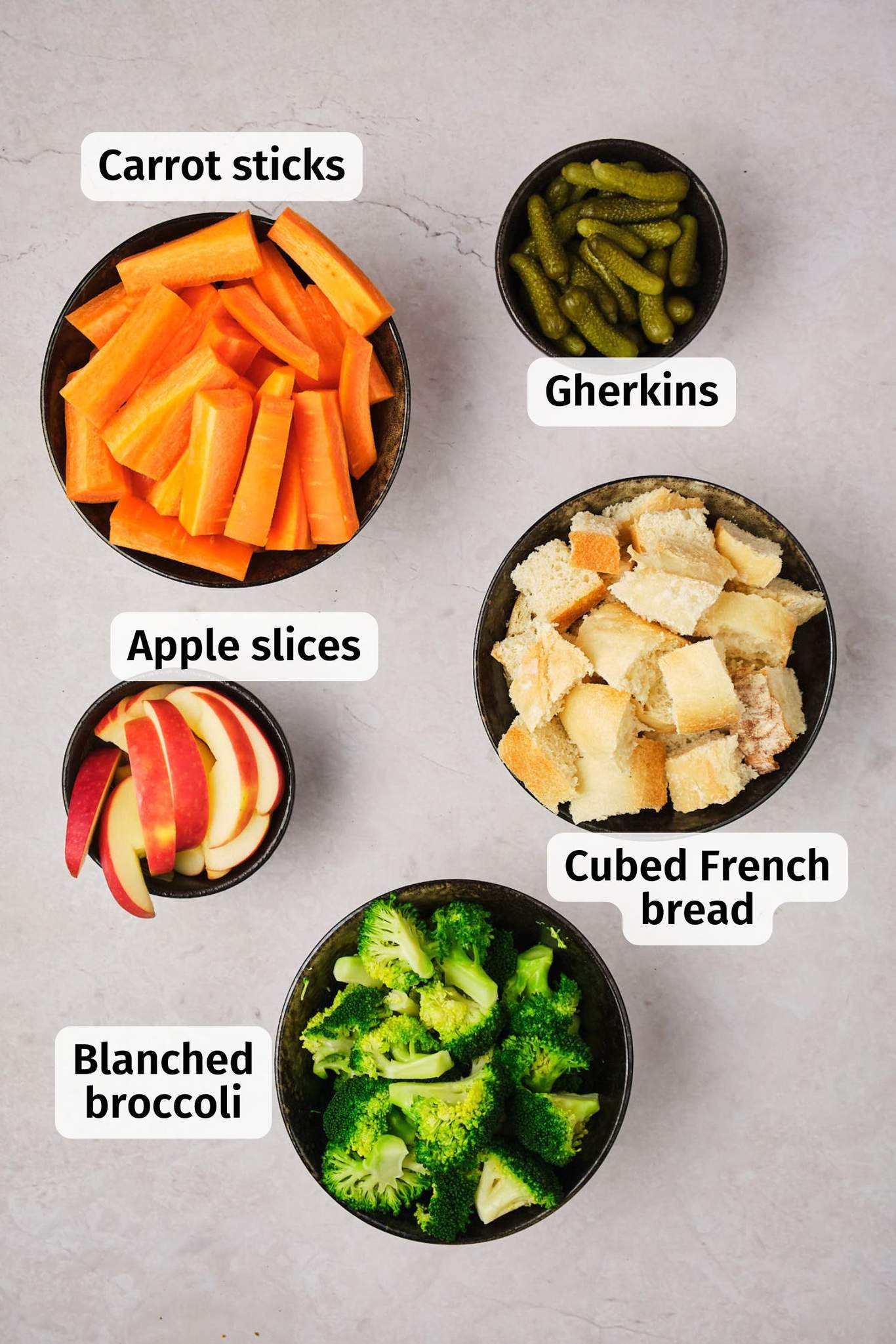 Items to be dipped in vegan fondue (apples, carrots, gherkins, broccoli, and bread), in individual bowls on a table, labeled. 