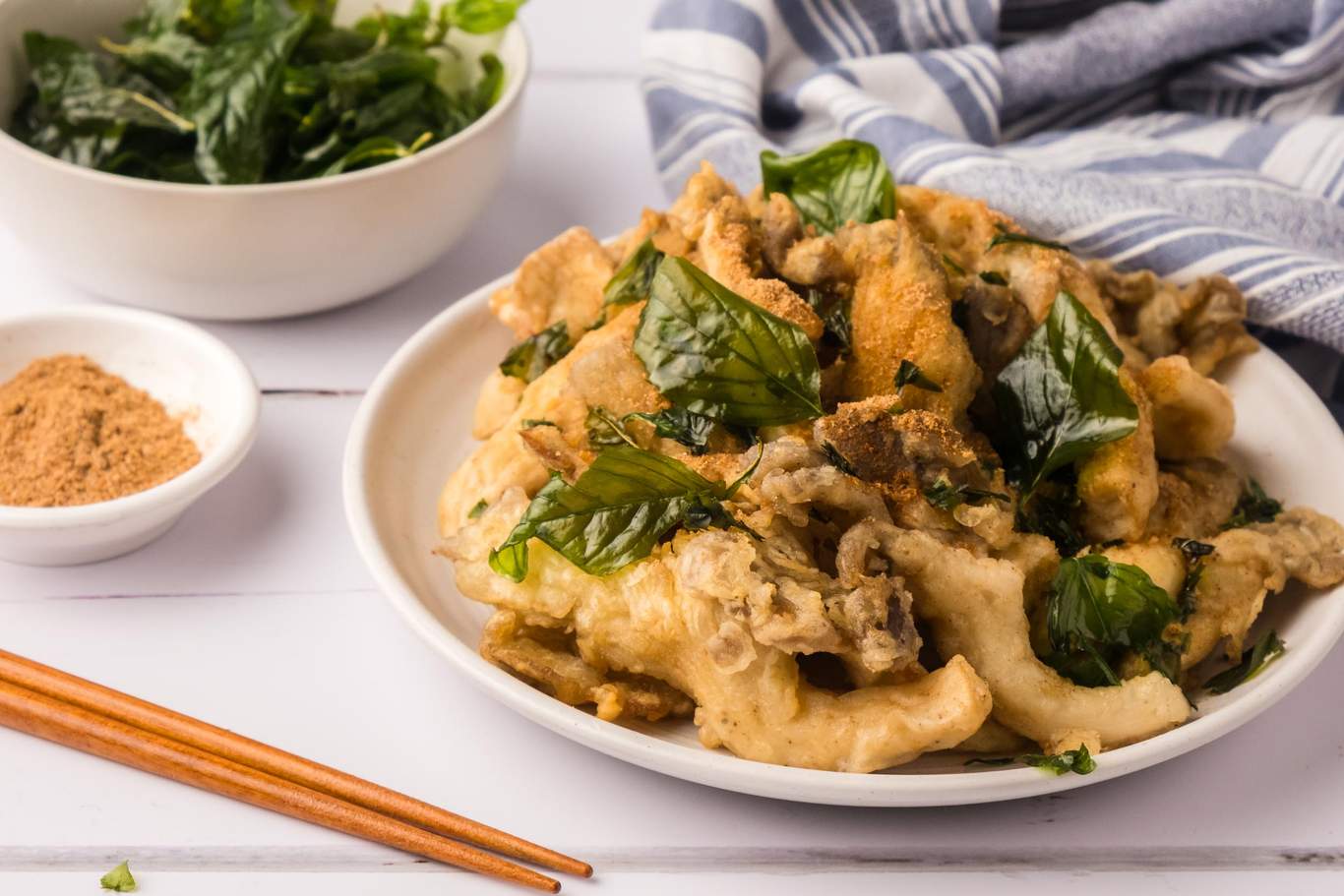Fried oyster mushrooms on a plate with fried basil, seasoning powder, and chopsticks to the side.