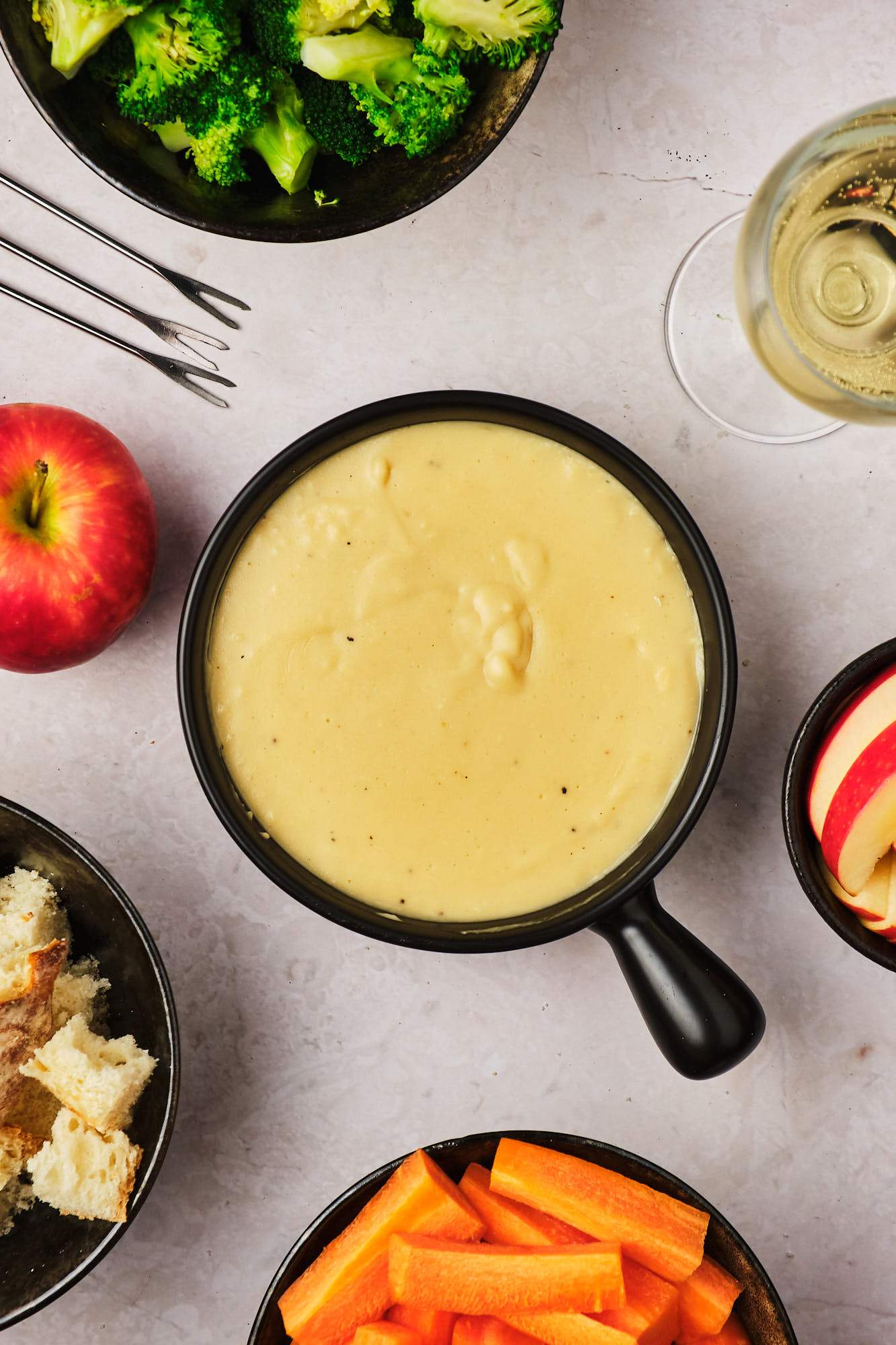 A pot of vegan fondue surrounded by bowls of fruits and vegetables, plus three fondue forks. 