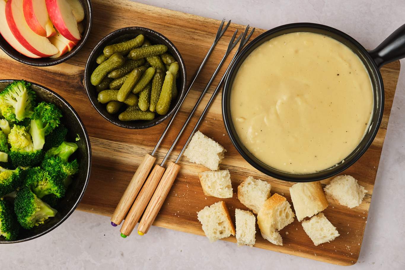 A fondue platter on a wooden serving board with a pot of vegan fondue, bread cubes, fondue forks, a bowl of gherkins, a bowl of broccoli, and a bowl of apple slices. 